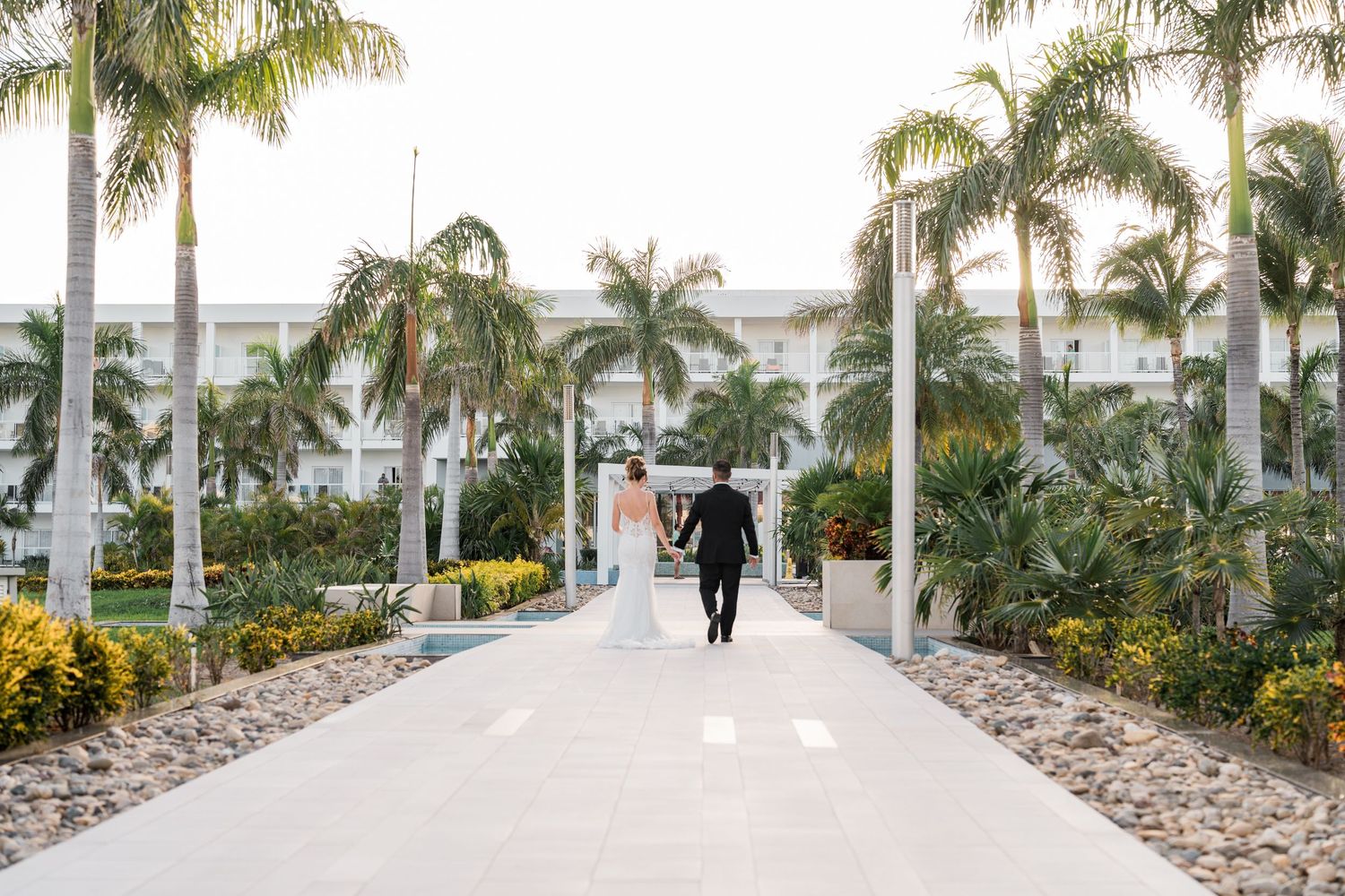 Bride and groom walk hand in hand down a palm-lined pathway at a tropical resort.