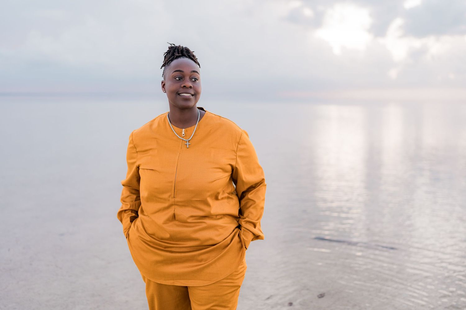 Smiling Black man in mustard yellow traditional outfit standing on beach with ocean backdrop.