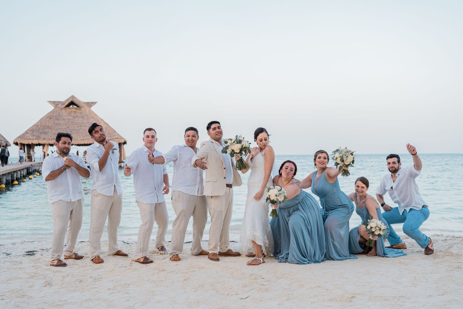 Wedding party celebrates with fun poses on tropical beach with overwater palapa at sunset.