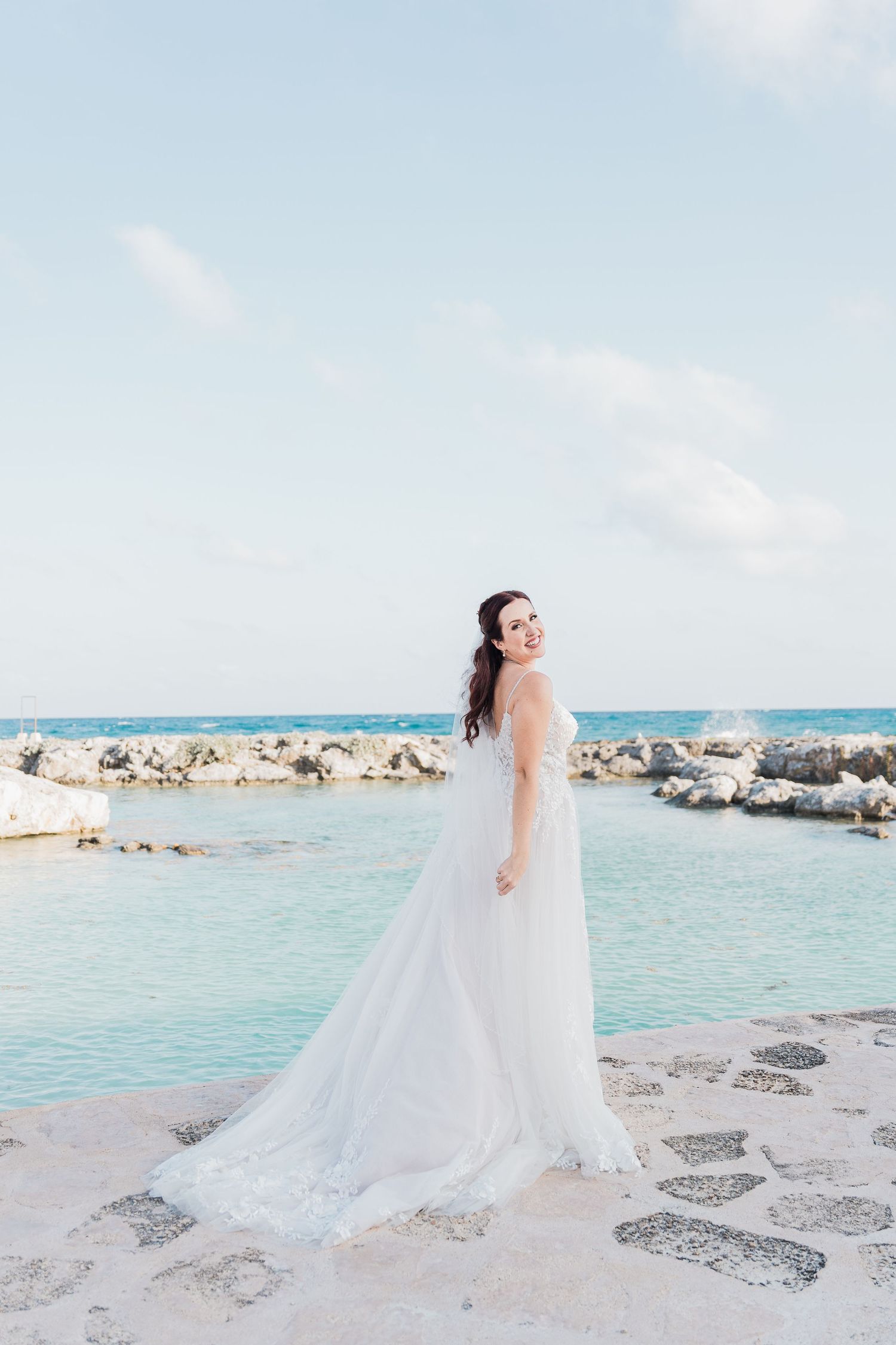 Smiling bride in a flowing white gown stands on a stone terrace with turquoise ocean waters in the background.
