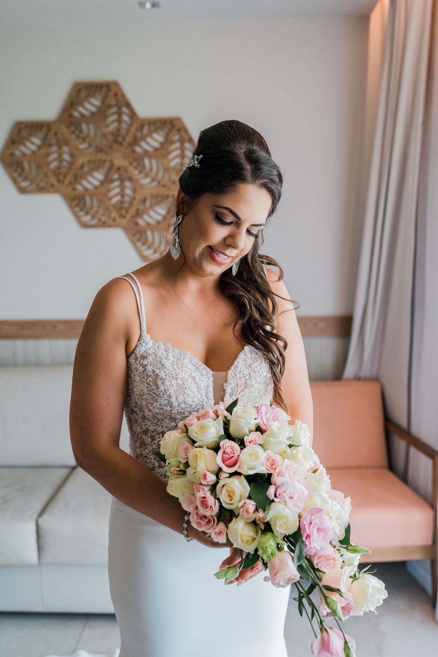 Smiling bride in beaded wedding gown admiring pink and white rose bouquet indoors.