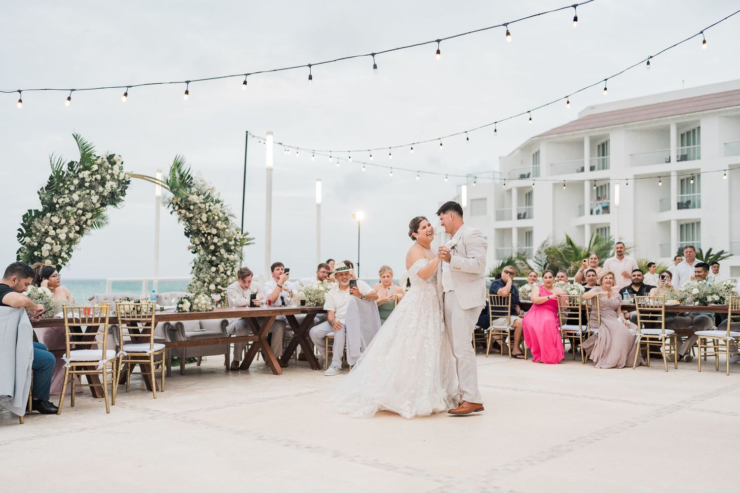 Bride and groom share first dance at outdoor beach wedding reception with string lights and floral arch.