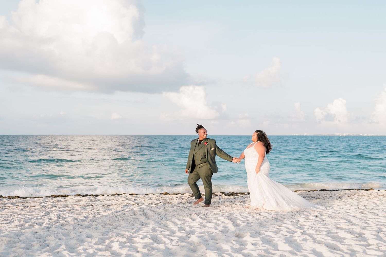 Newlywed couple in wedding attire walks hand in hand on a white sandy tropical beach by turquoise water.