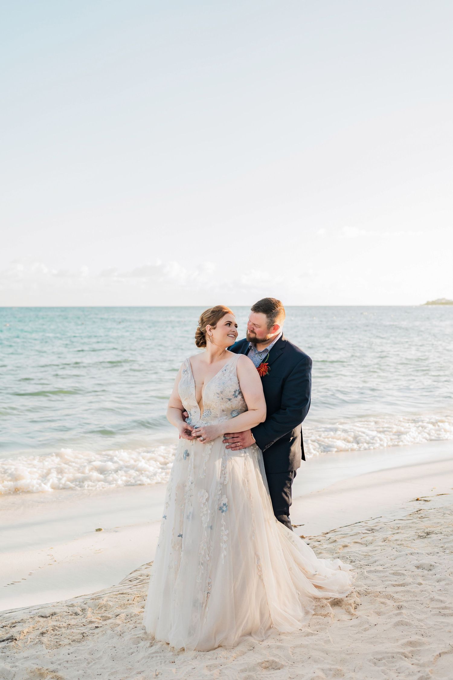 Secrets Akumal Wedding - A bride and groom embrace on a sandy beach at sunset, with calm turquoise ocean waves behind them.