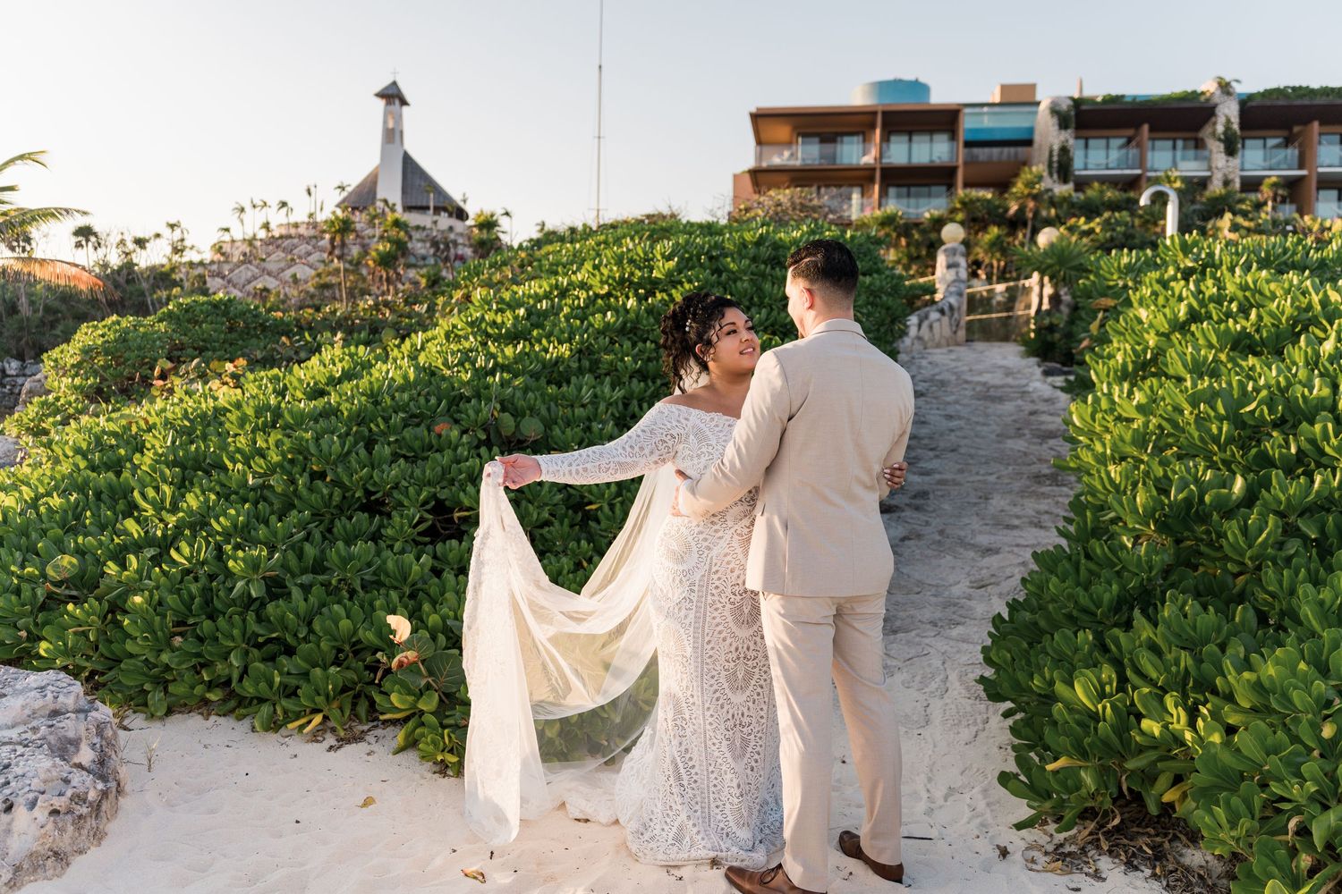 Xcaret Arte Wedding - Bride and groom walking on sandy beach path surrounded by lush greenery with resort buildings in background.