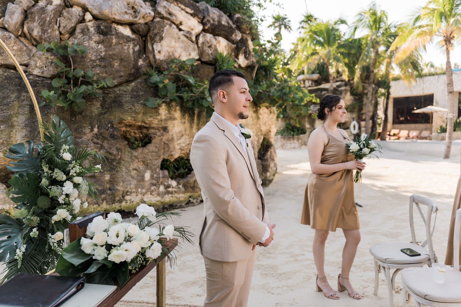 Xcaret Arte Wedding - Groom in beige suit stands at outdoor beach wedding ceremony altar decorated with white flowers and greenery.