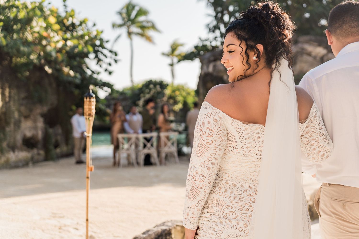 Xcaret Arte Wedding - Bride in lace off-shoulder gown with veil at tropical beach wedding ceremony with tiki torch.