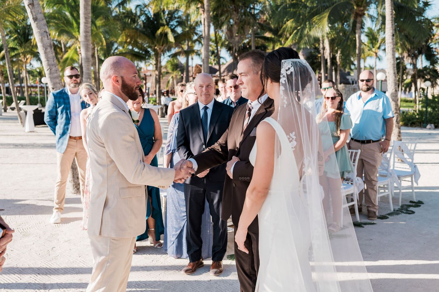 Couple exchanging vows at an outdoor beach wedding ceremony surrounded by palm trees and seated guests.