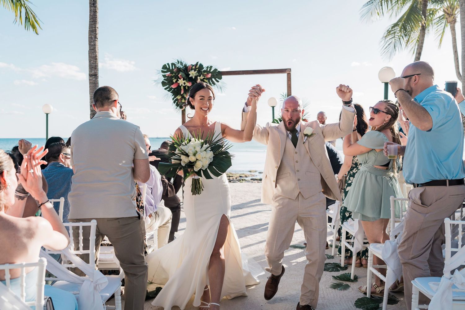Joyful newlyweds celebrate at outdoor beach wedding ceremony, bride in white gown with floral crown, groom in tan suit.