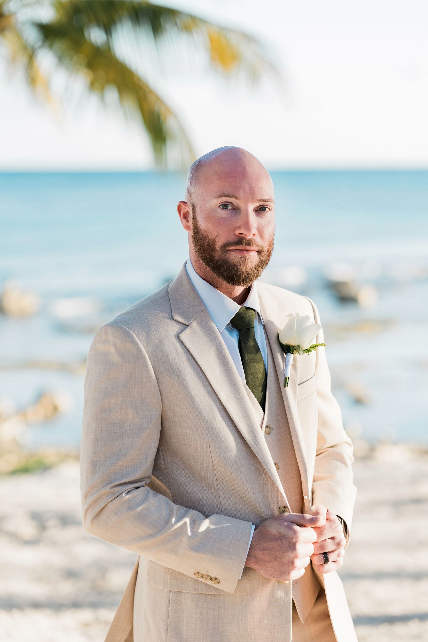 Bald groom with beard in tan suit and green tie wearing white boutonniere poses on tropical beach.