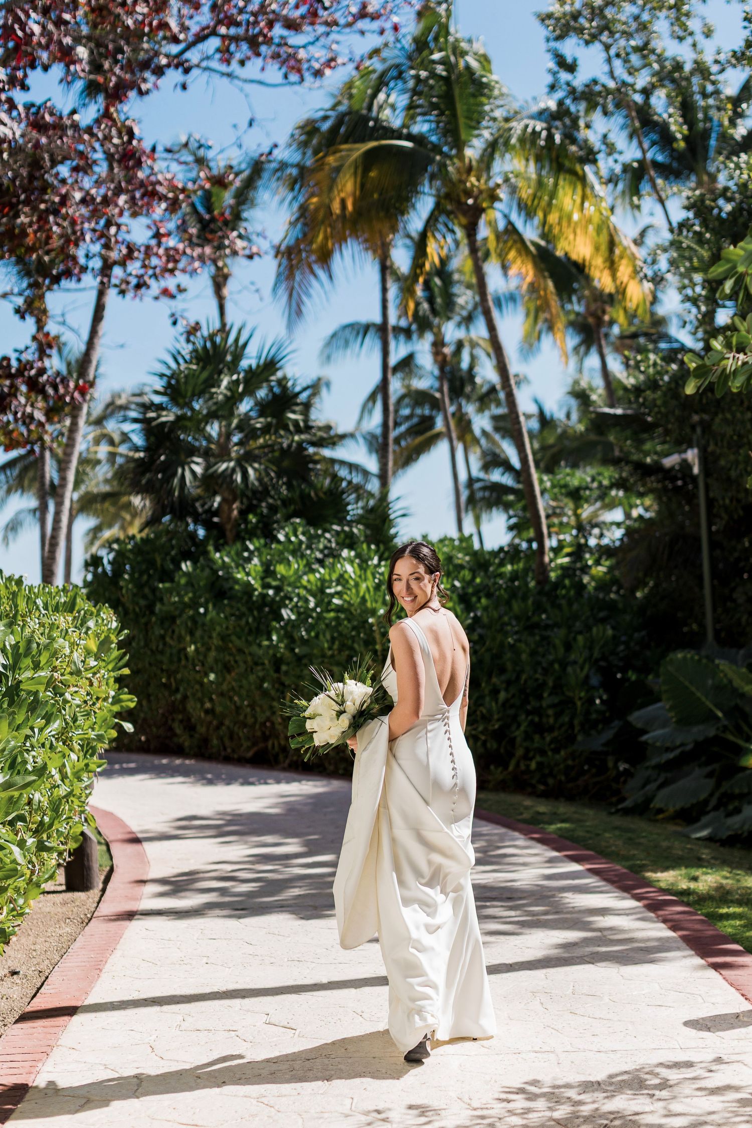 Smiling bride in backless white gown holding bouquet walks tropical garden path with palm trees.