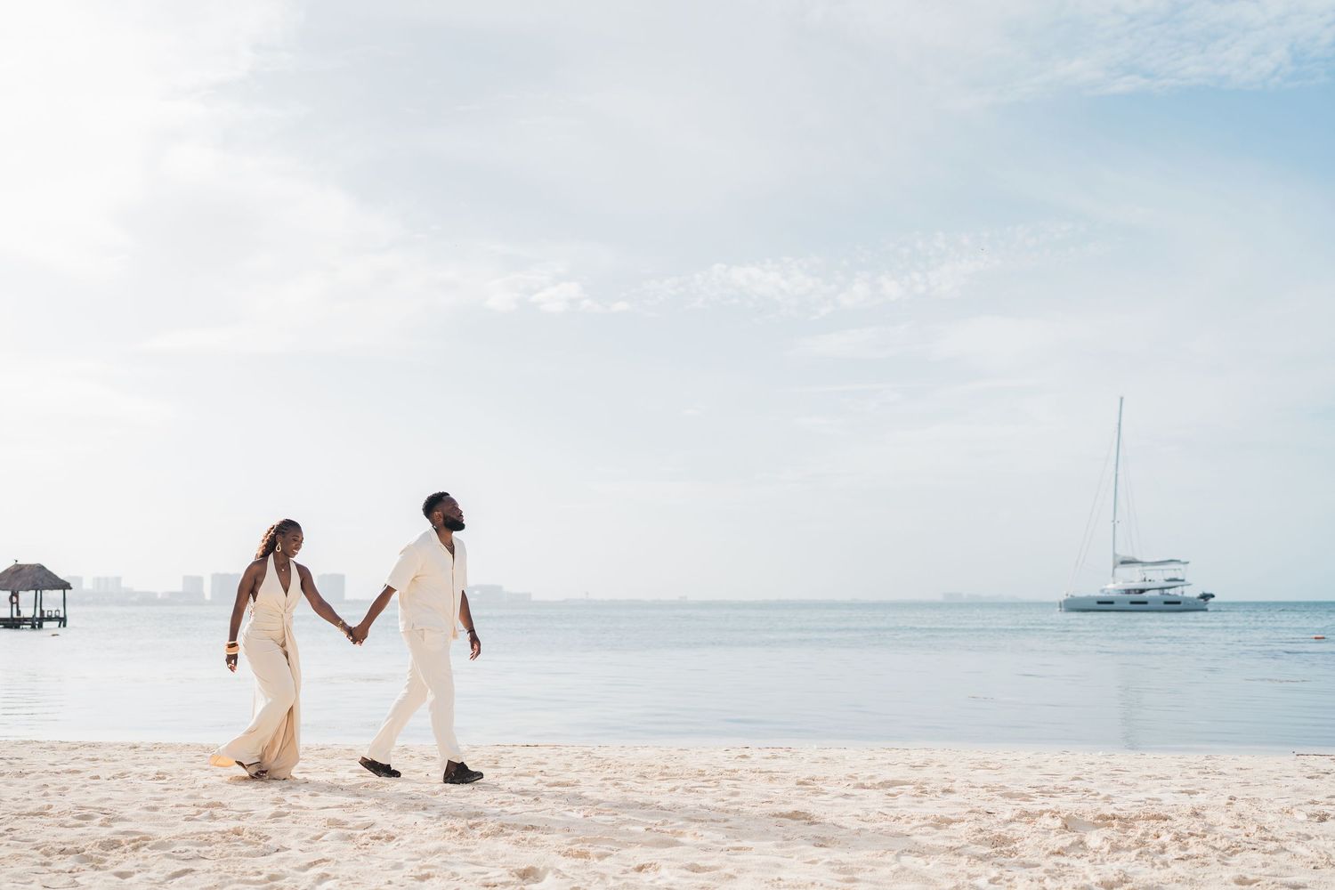 Couple in white attire holding hands walking on a tropical beach with a sailboat in the background.