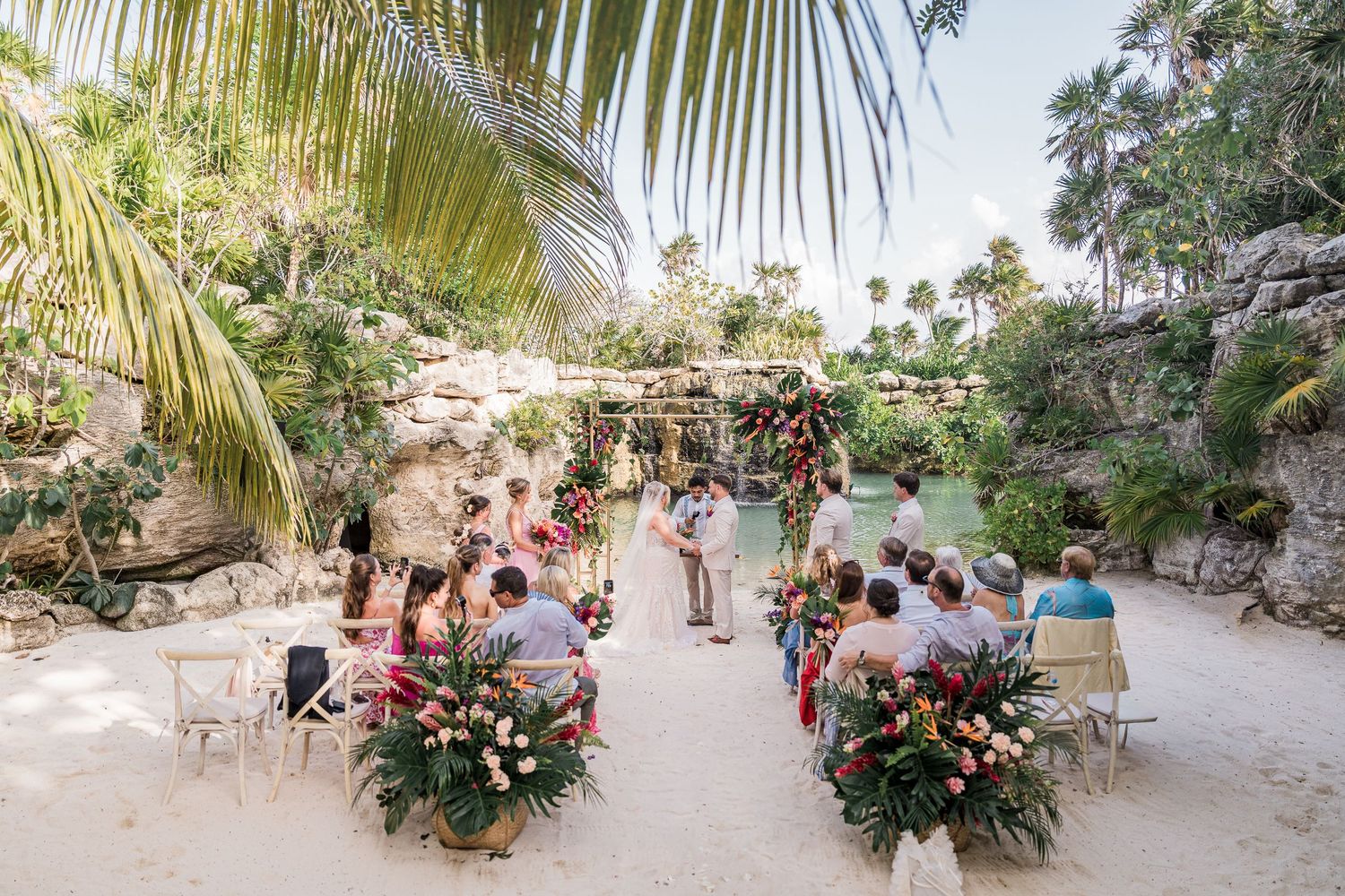 Tropical beach wedding ceremony with guests seated on white chairs surrounded by lush palms and rocky cliffs.