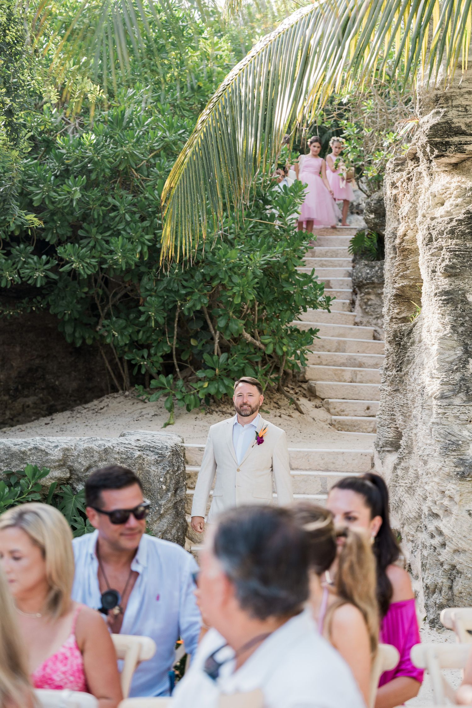 Groom in beige suit awaits bridesmaids in pink dresses descending stone steps at tropical outdoor wedding venue.