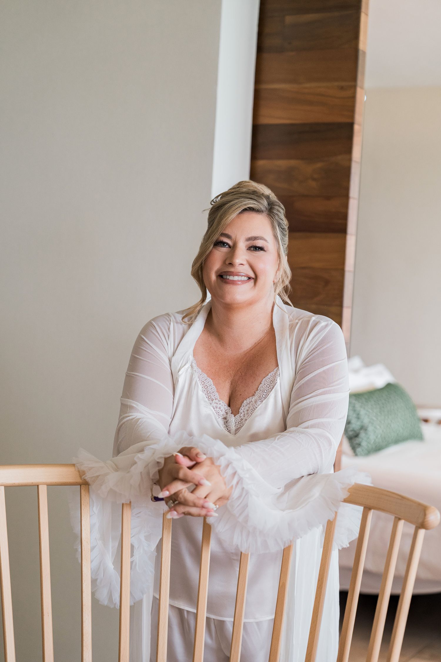 Smiling bride in white lace robe leaning on wooden railing in bright bedroom on wedding morning.