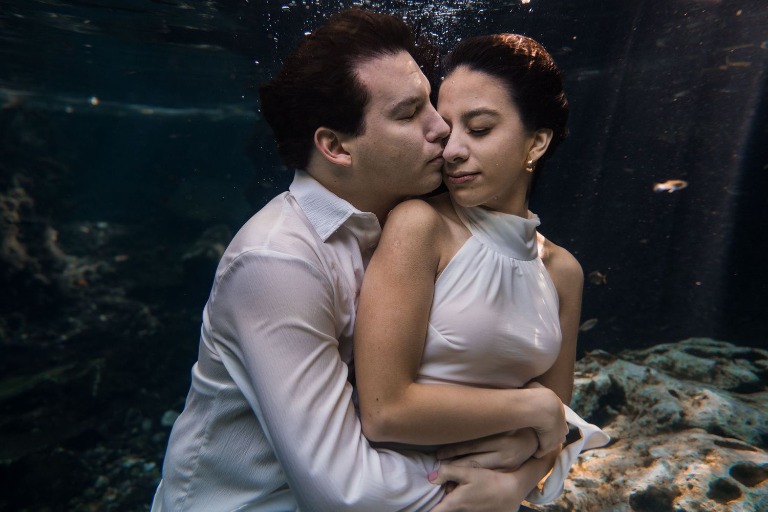 Romantic couple embracing underwater in a cenote, wearing white outfits, surrounded by fish and rock formations.