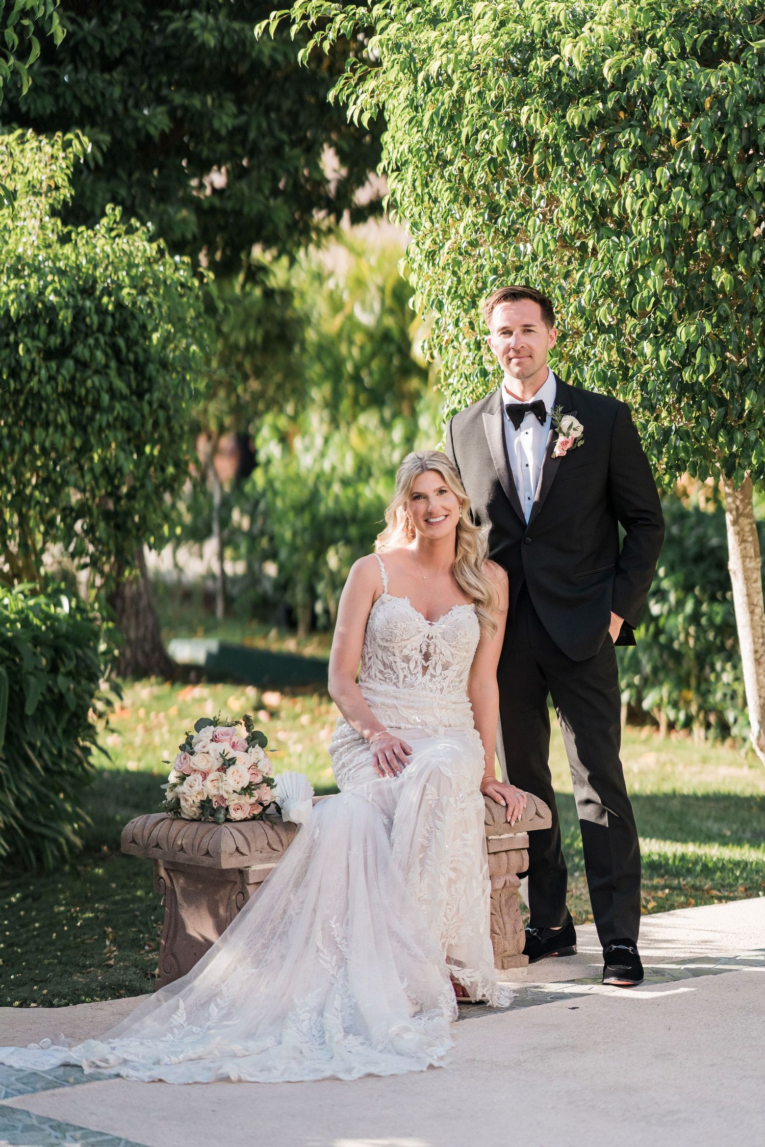 Bride in lace gown and groom in tuxedo pose together on stone bench surrounded by lush greenery on their wedding day.