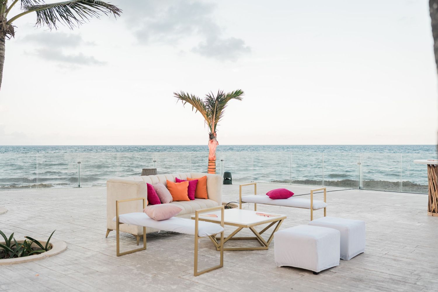 Elegant beachfront lounge area with white and gold furniture, colorful pillows, and a palm tree by the ocean.