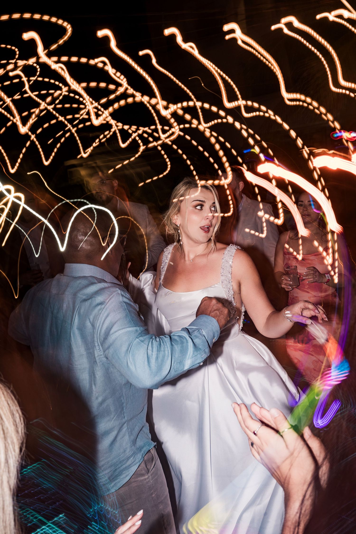 Bride in white gown dancing with guests at a wedding reception, surrounded by colorful light trails at night.
