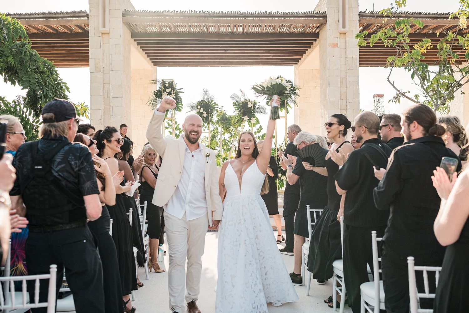Joyful newlyweds walk down the aisle at an outdoor wedding ceremony as guests in black attire celebrate.