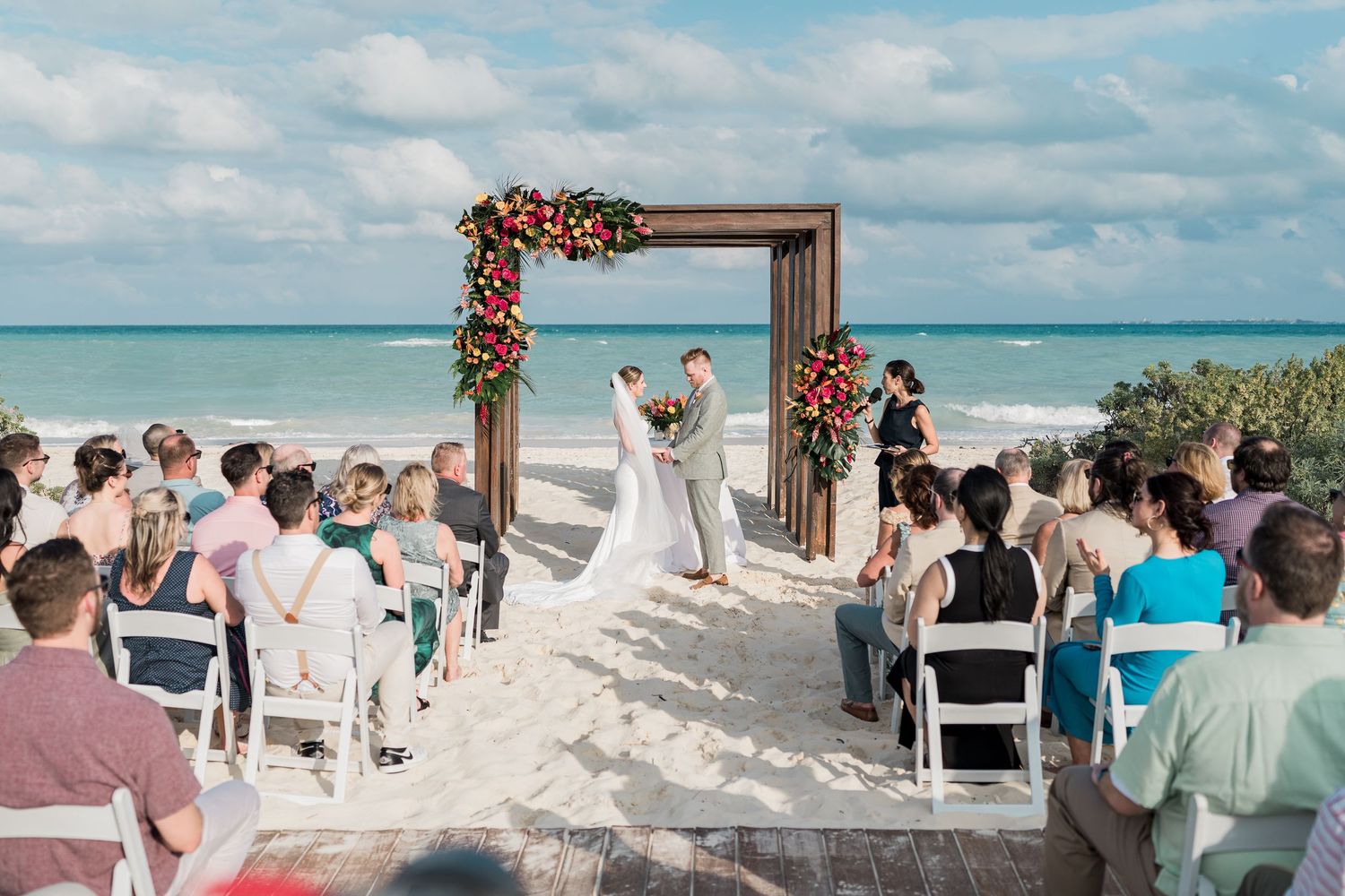 Beach wedding ceremony with floral arch, bride and groom, guests seated on white chairs, ocean backdrop.