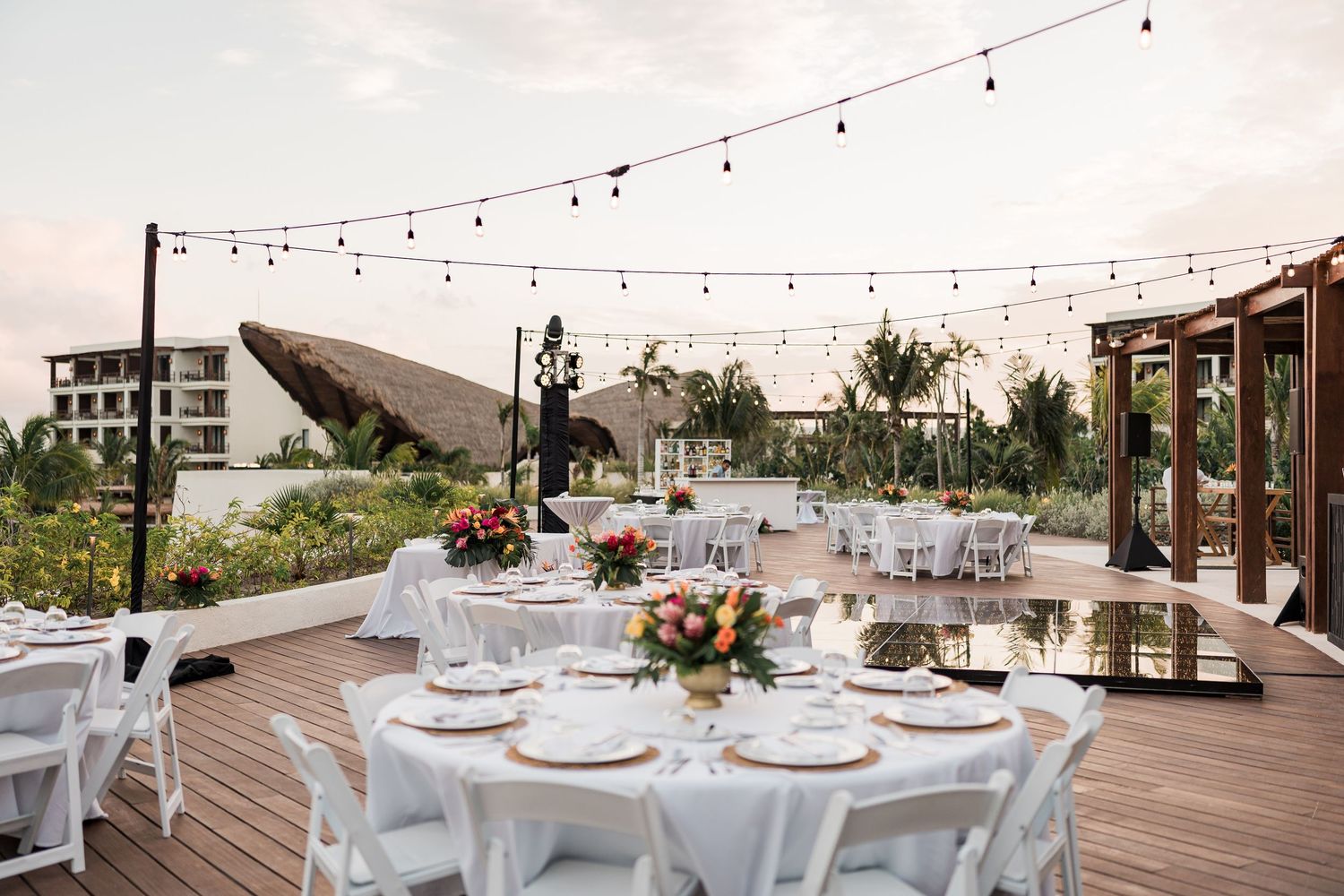 Elegant outdoor rooftop wedding reception with white round tables, string lights, and colorful floral centerpieces at sunset.