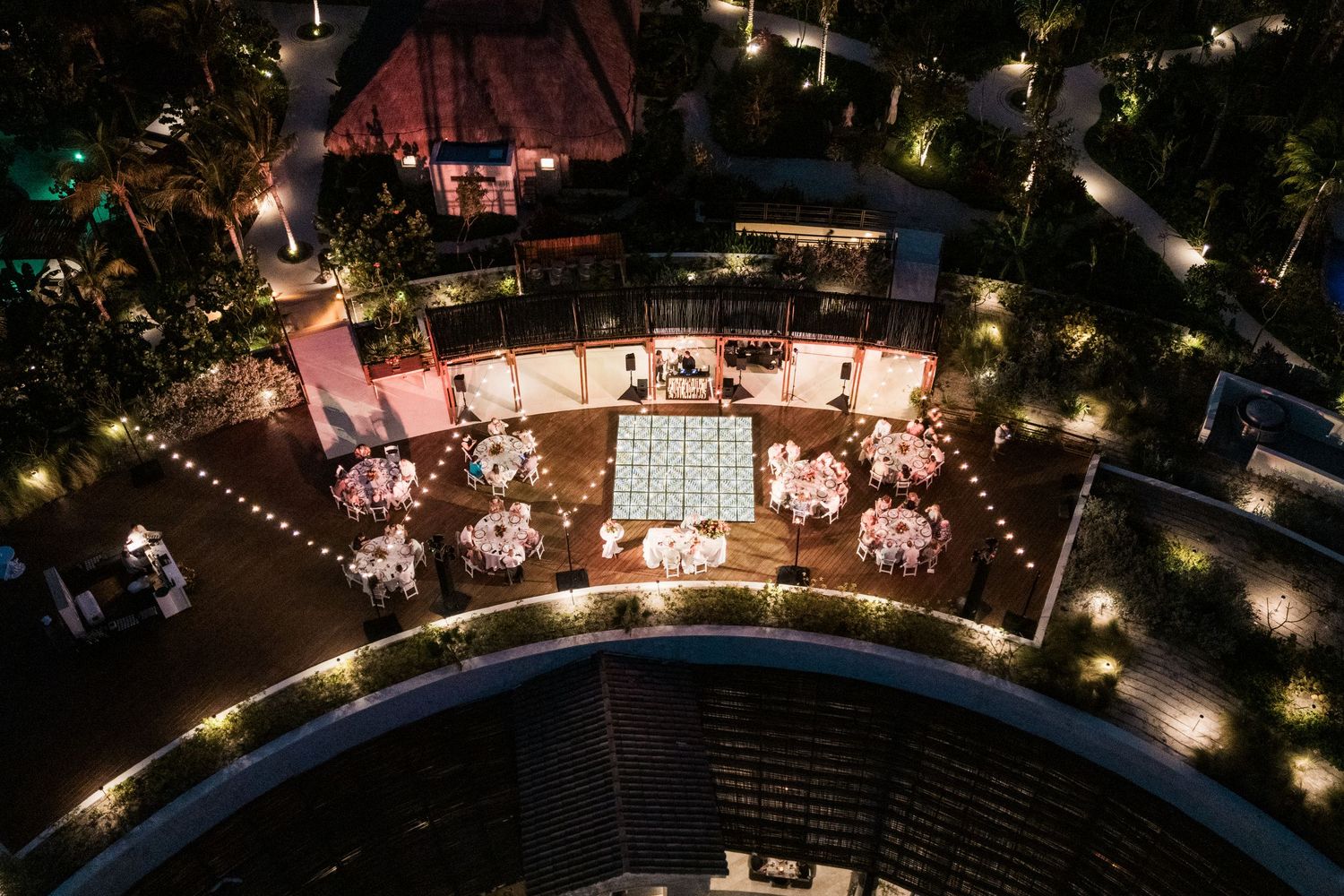 Aerial night view of an elegant outdoor wedding reception with string lights, round tables, and a dance floor.