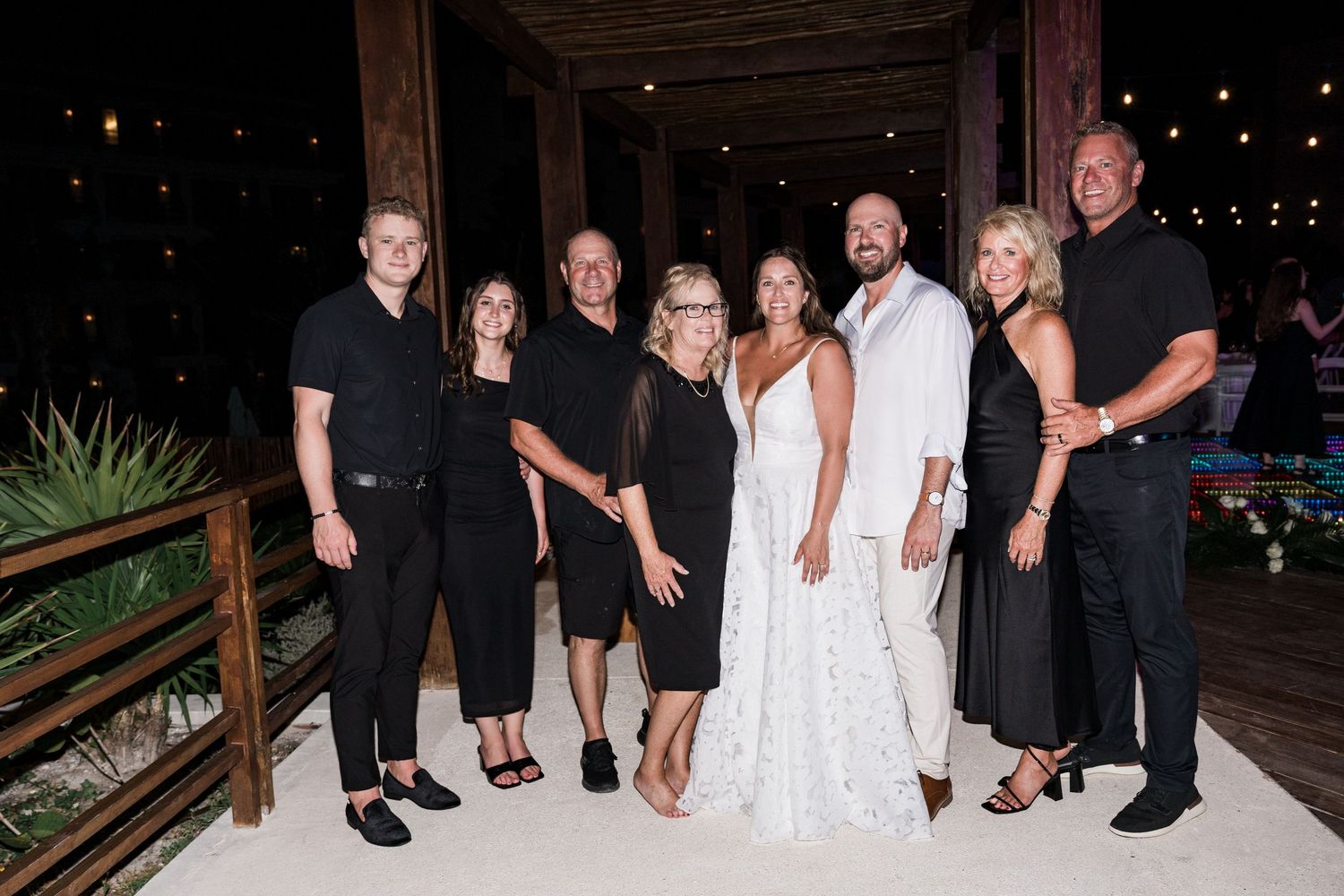 Group of eight people in formal black and white attire posing together at an outdoor evening wedding celebration.