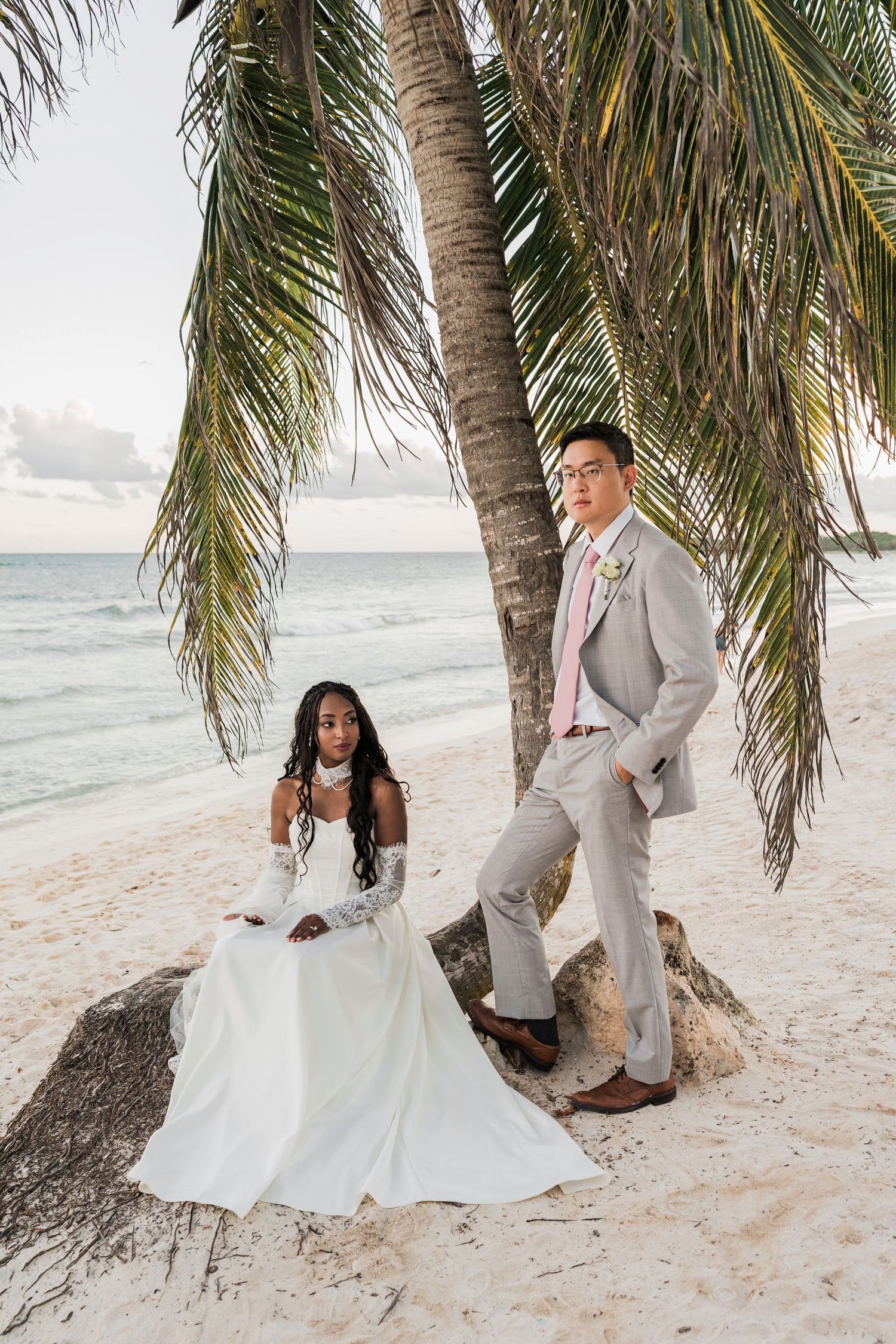 Bride and groom pose under a palm tree on a sandy beach, she in a white gown, he in a gray suit with pink tie. Wedding at Unico 2087