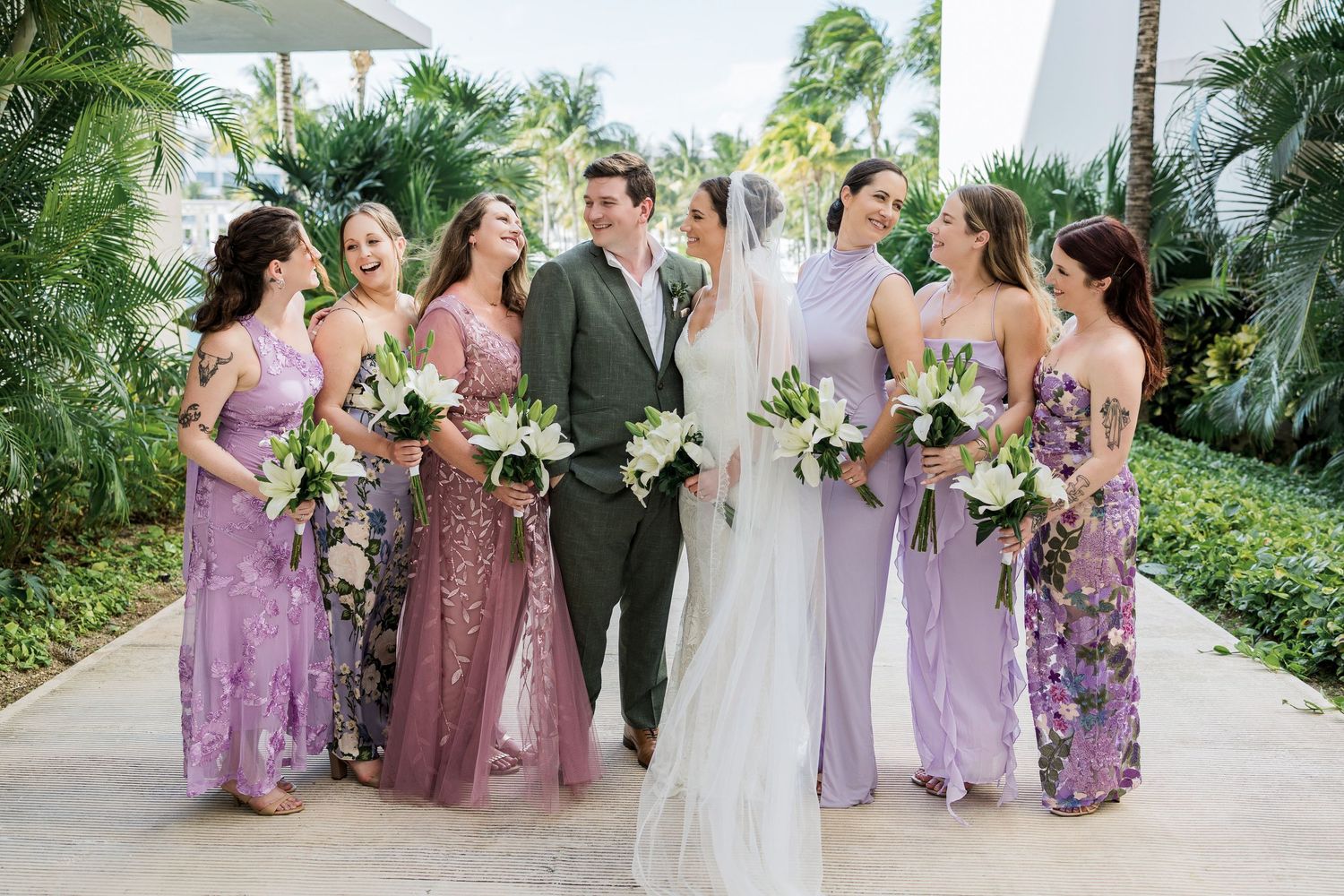 Bride and groom walk with bridesmaids in mismatched purple gowns holding white bouquets at tropical resort wedding.