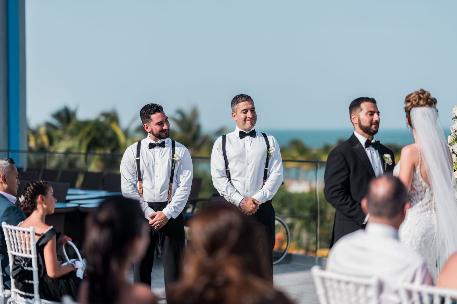 Smiling groomsmen in black tuxedos with suspenders stand beside the bride at an outdoor beach wedding.