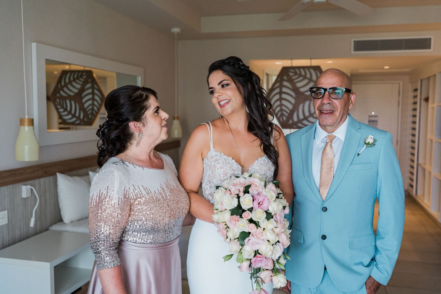Bride smiling with parents in hotel room, holding pink bouquet, father in light blue suit.