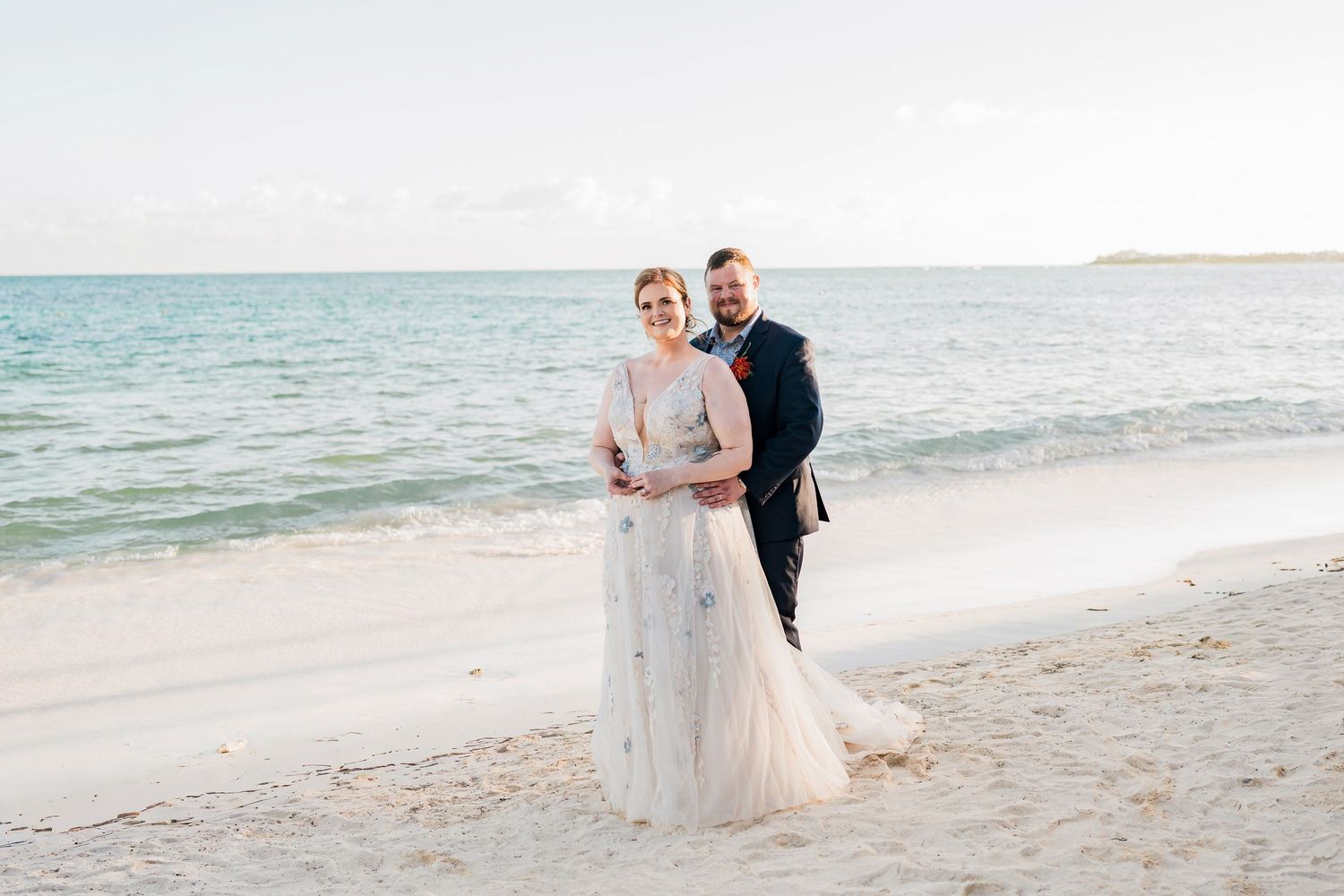 Secrets Akumal Wedding - Bride and groom smiling together on a sandy beach with calm ocean waves in the background on their wedding day.
