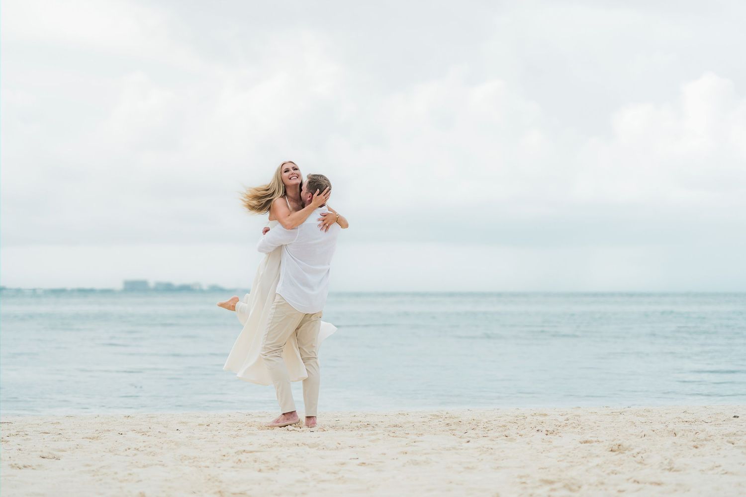 Cancun Engagement Photoshoot - Couple embracing joyfully on a sandy beach in white outfits with calm ocean waves in the background.