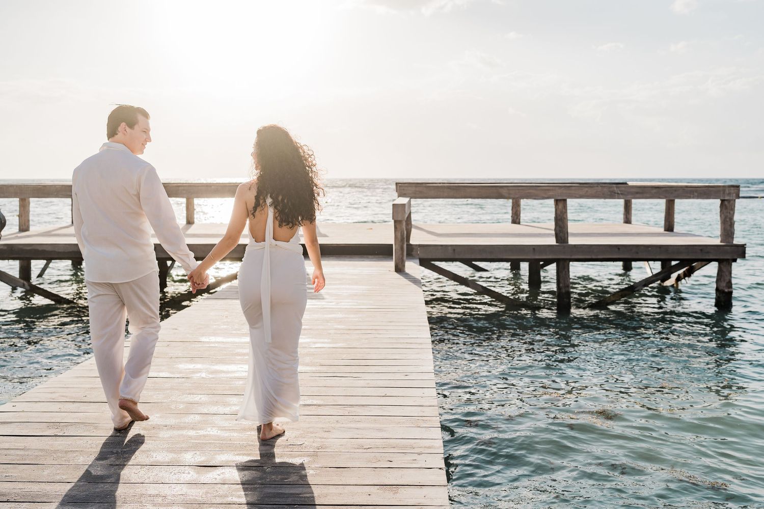 Couple in white outfits holding hands walking on a wooden pier over turquoise ocean water at sunset.