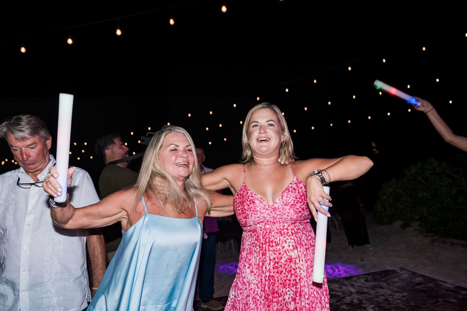 Hotel Xcaret México Two women smile and dance holding glow sticks at an outdoor evening wedding reception with string lights.