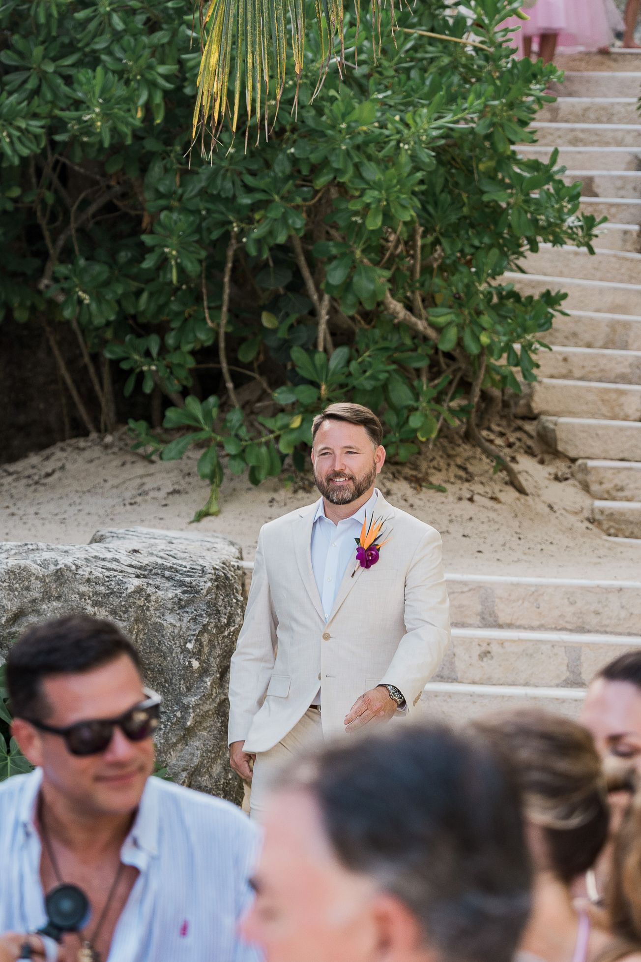 Hotel Xcaret México Wedding  - Groom in beige suit with tropical boutonniere smiling at outdoor beach wedding ceremony with stone steps.