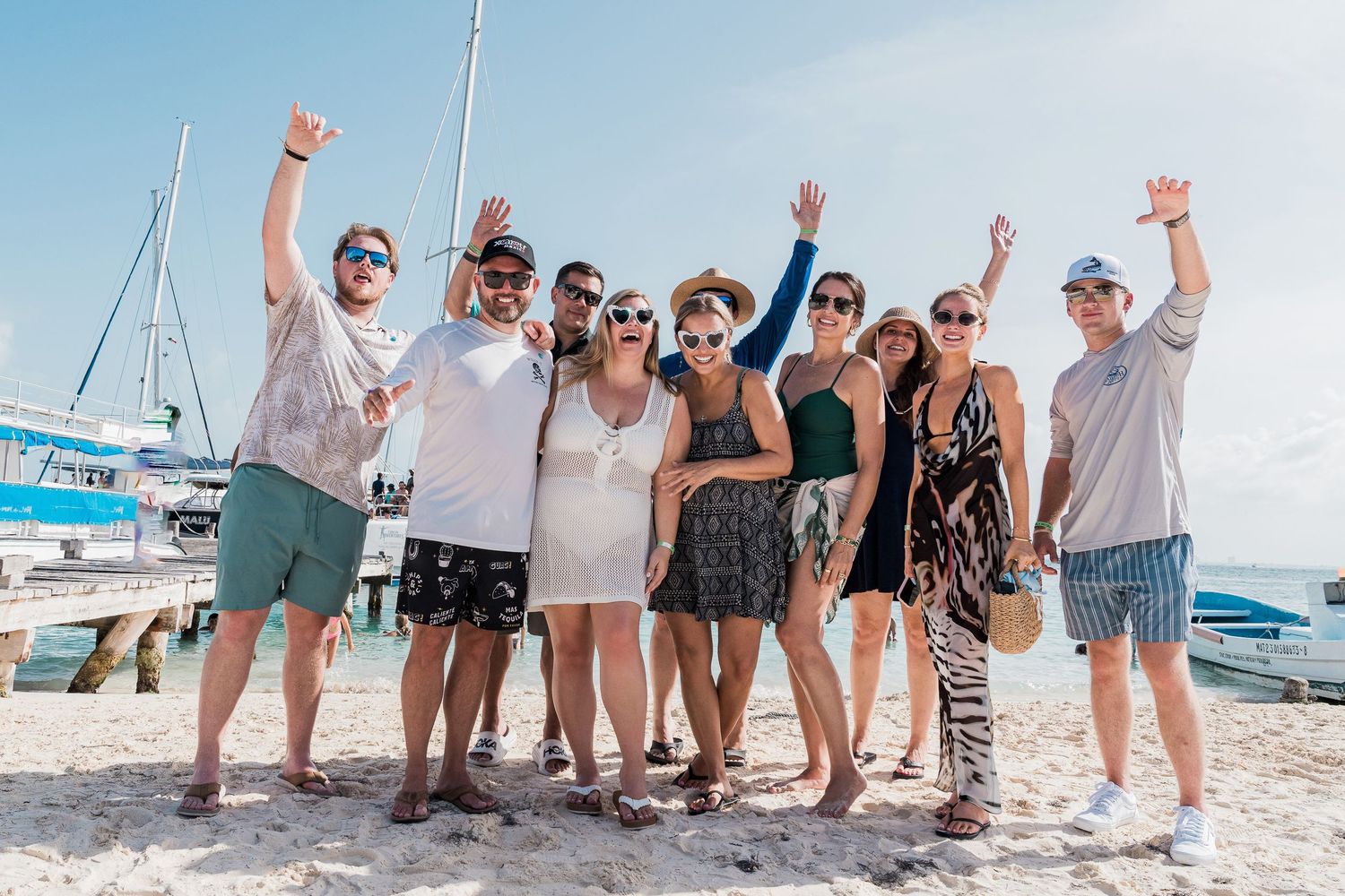 Riviera Maya Multi wedding day - Group of friends posing and celebrating on a sunny beach with sailboats in the background.