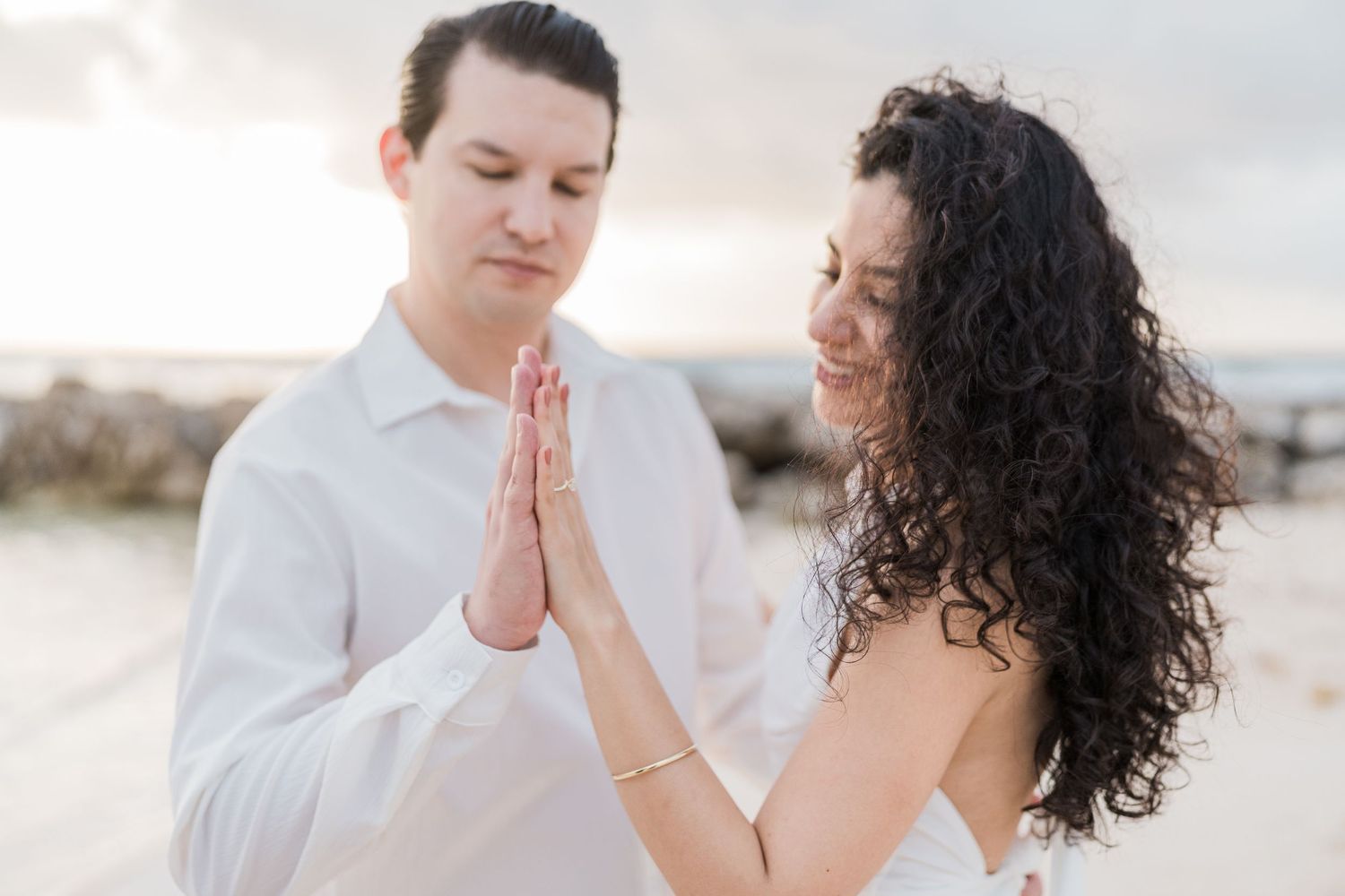 Couple in white attire practicing prayer hands pose together on a beach at sunset.