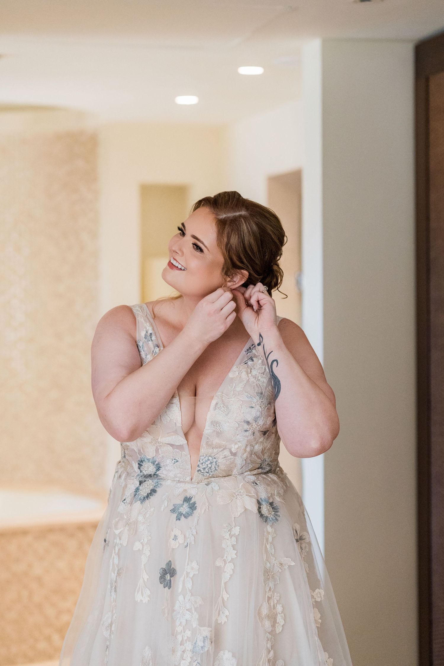 Bride in floral wedding gown putting on earrings while getting ready, smiling in a bright room.