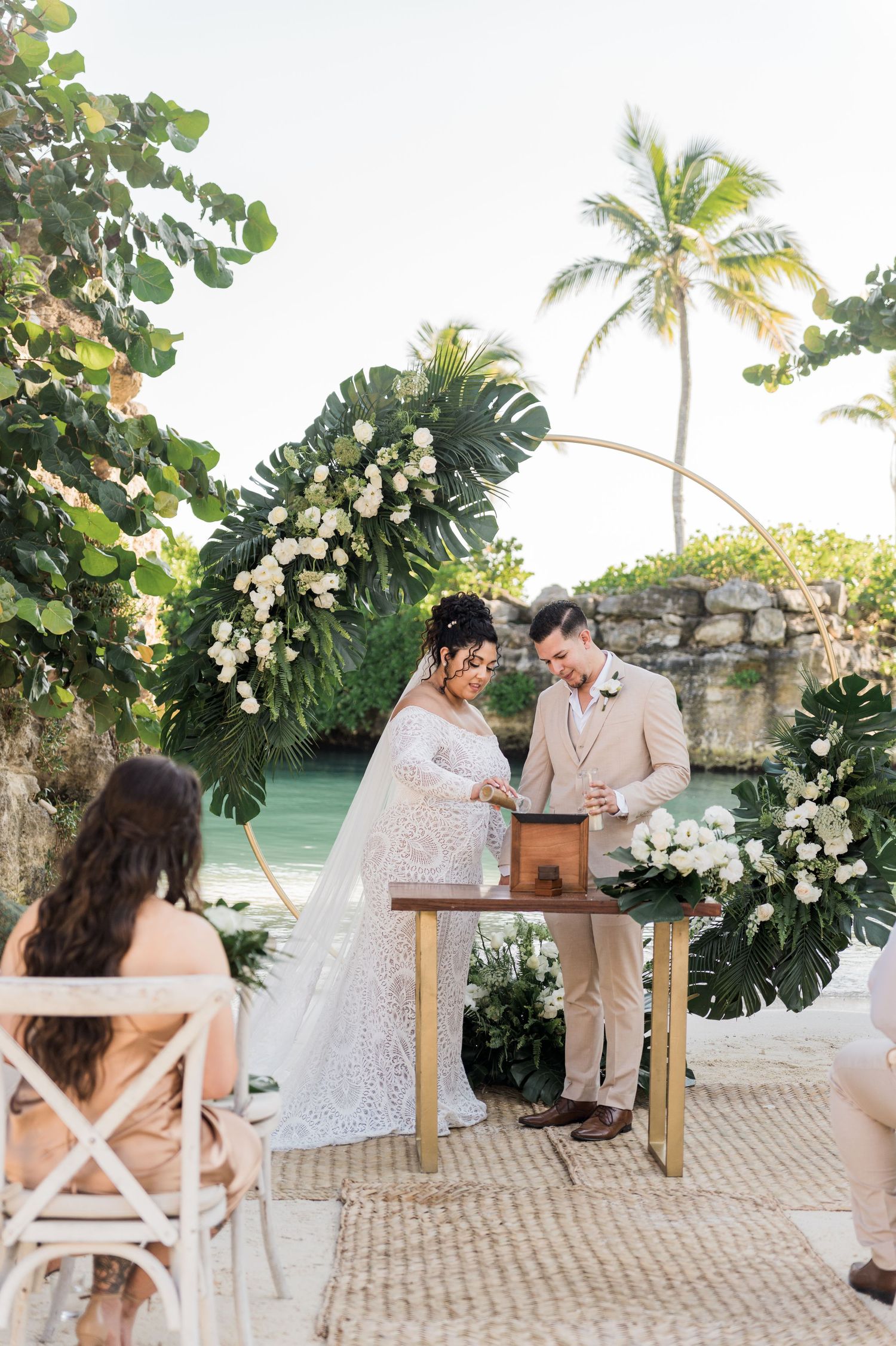 Xcaret Arte Wedding - Bride and groom at tropical beach wedding ceremony with white floral arch, palm trees, and turquoise water backdrop.