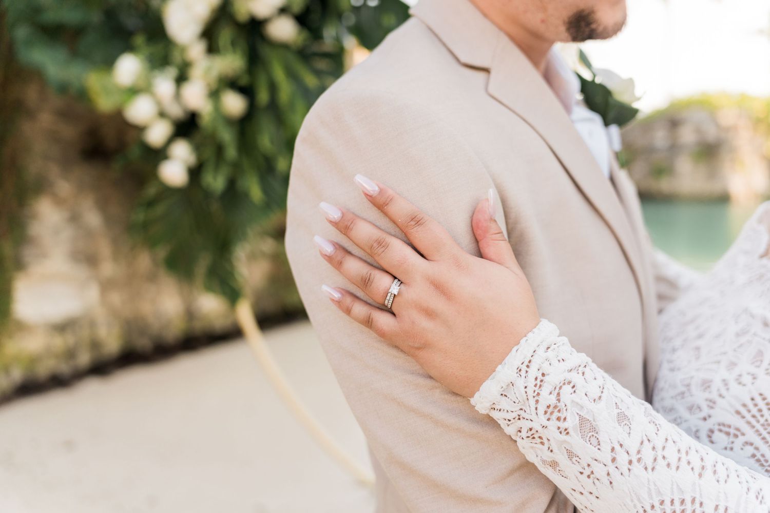 Xcaret Arte Wedding - Bride's hand with diamond ring resting on groom's beige suit shoulder at outdoor wedding ceremony.
