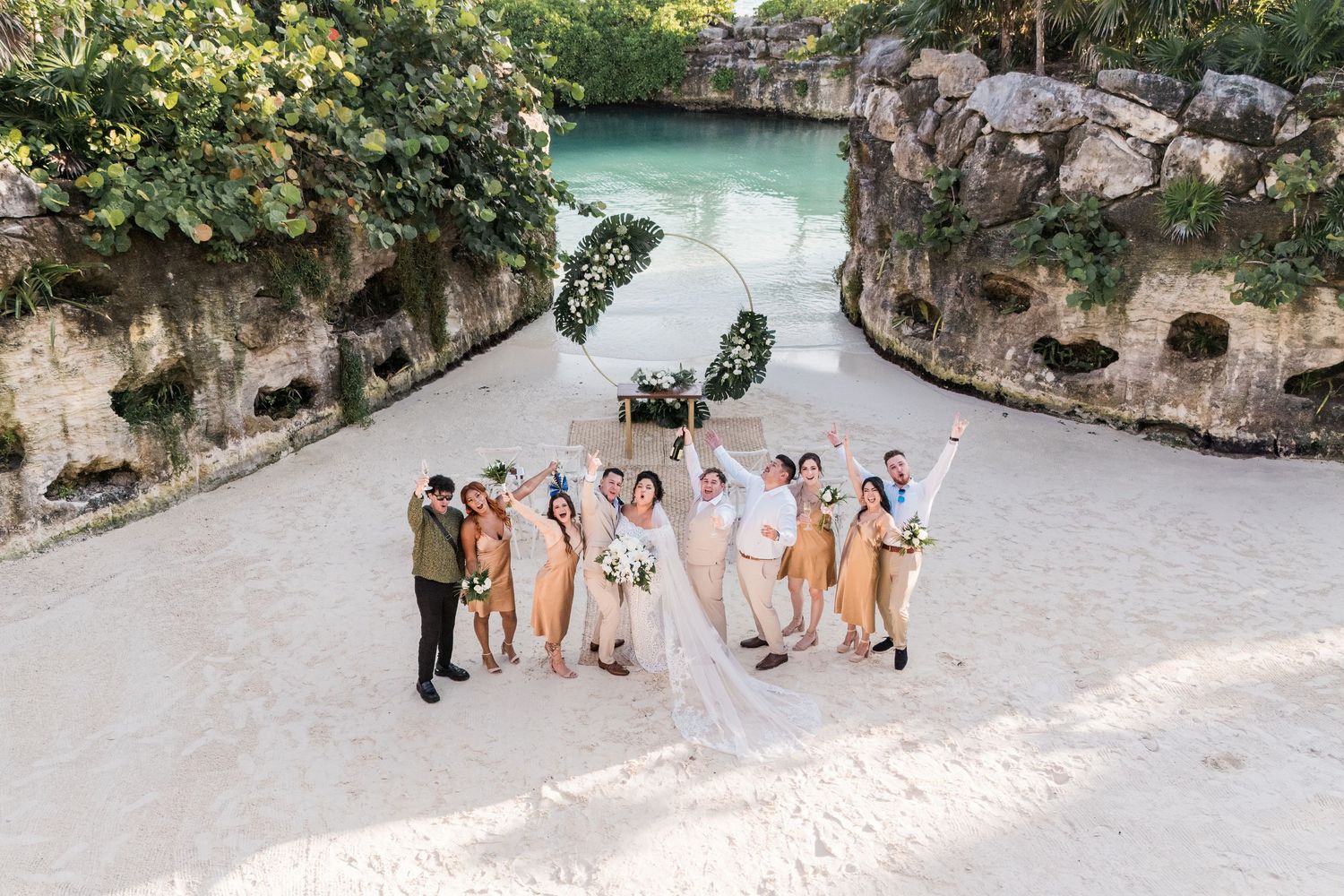 Xcaret Arte Wedding - Wedding party poses on white sand beach by turquoise cenote with circular floral arch backdrop in Mexico.