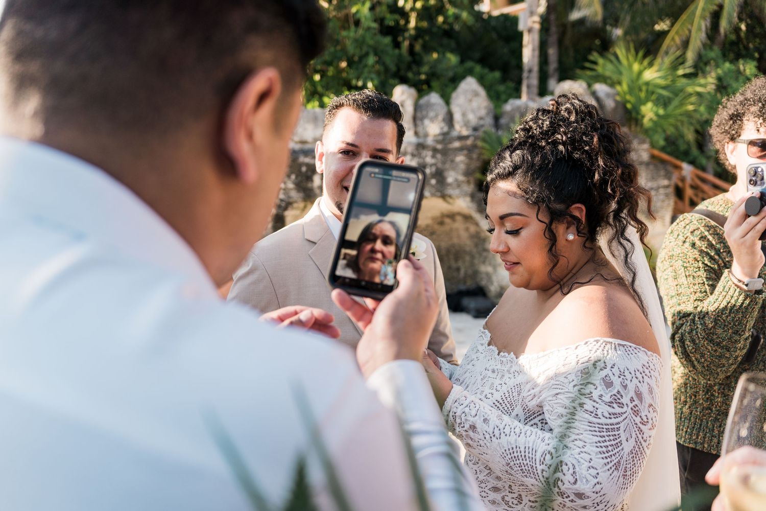 Xcaret Arte Wedding - Bride and groom share wedding vows while a guest video calls a loved one on a smartphone during an outdoor ceremony.