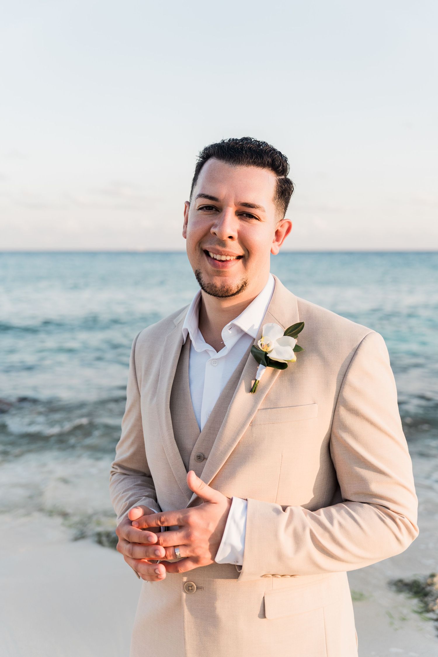 Xcaret Arte Wedding - Groom in beige suit with white boutonniere smiling on sandy beach with turquoise ocean backdrop.
