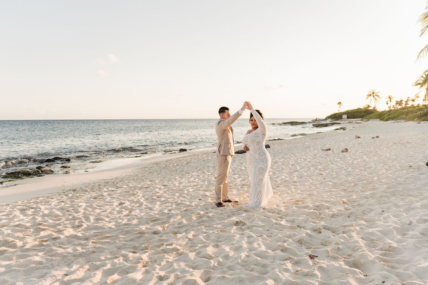 Xcaret Arte Wedding - Couple dancing on a white sand beach at sunset in white wedding attire with ocean and palm trees in background.
