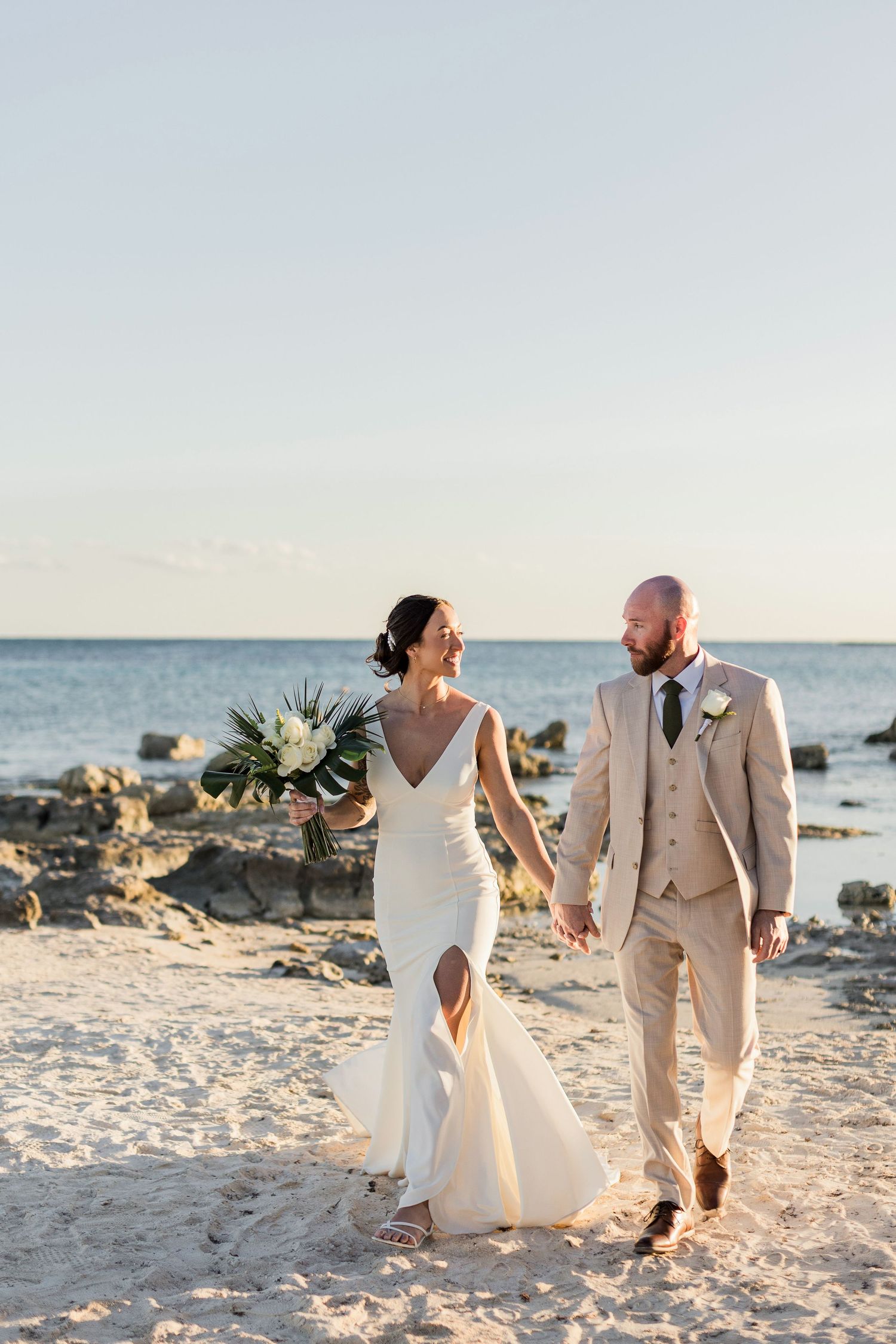 Bride and groom walk hand-in-hand on a sandy beach at sunset, she in a white gown, he in a tan suit.