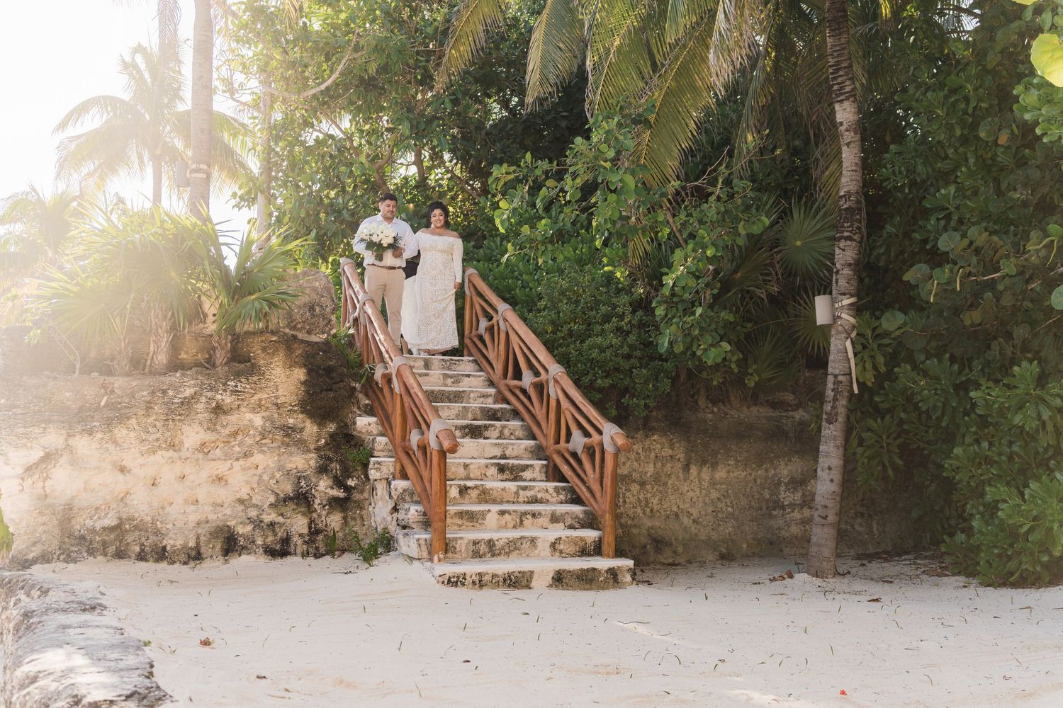 Xcaret Arte Wedding - Couple in wedding attire descending tropical beach stairs surrounded by lush palm trees in golden sunset light.