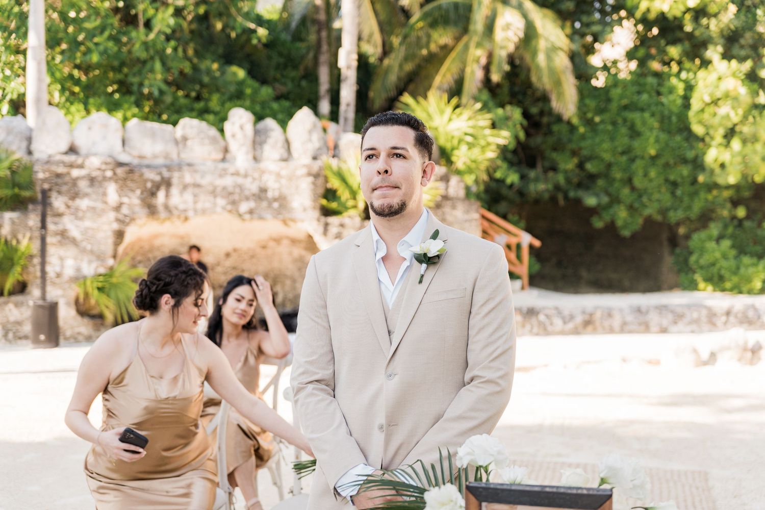 Xcaret Arte Wedding - Groom in beige suit stands at outdoor wedding altar with bridesmaids in gold dresses in tropical setting.