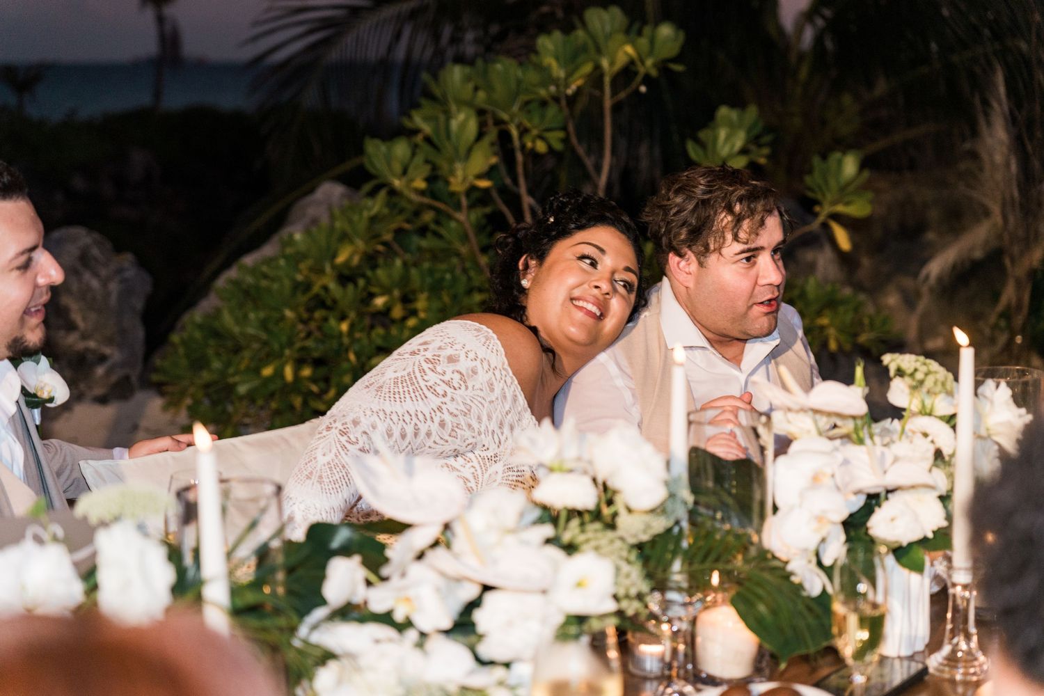 Xcaret Arte Wedding - Bride and groom laughing at outdoor wedding reception table adorned with white roses and candles at dusk.